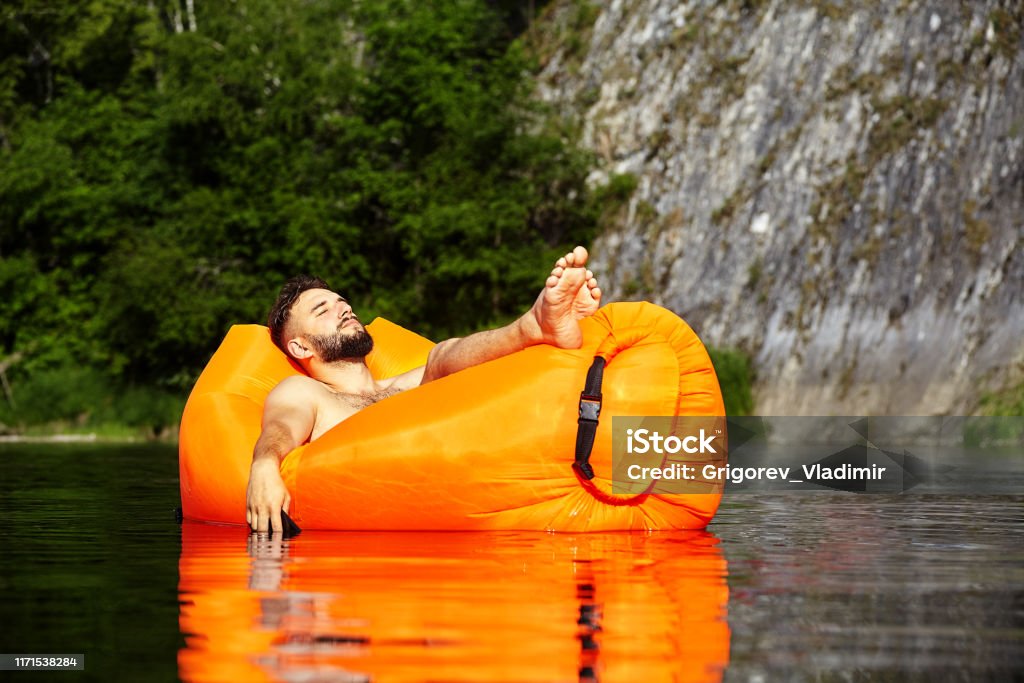 Man lying on an orange inflatable lounger in a body of water with trees and rocks in the background.