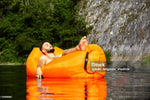 Load image into Gallery viewer, Man lying on an orange inflatable lounger in a body of water with trees and rocks in the background.
